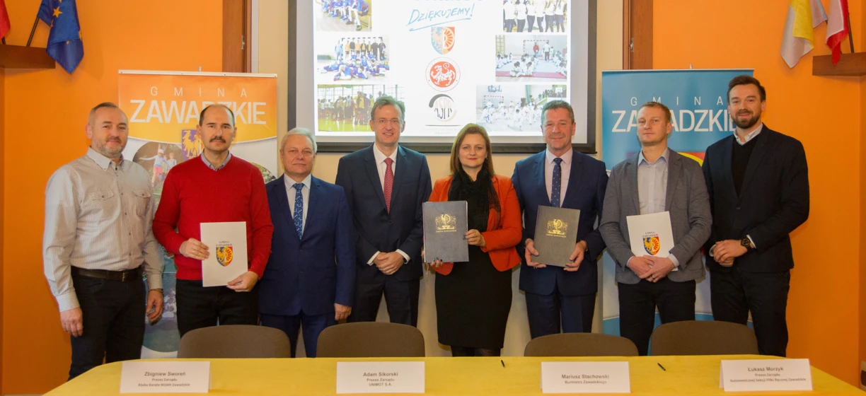 Image alt-text: Group of white-collar workers in formal wear, including suits, blazers, and neckties, standing and smiling. Logo with the text "GMINA" visible.