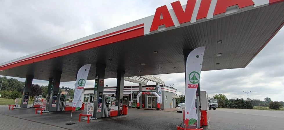 Image of a car parked at a filling station with the fuel pump nozzle in the gas tank. The vehicle's mirrors, bumper, and registration plate are visible.