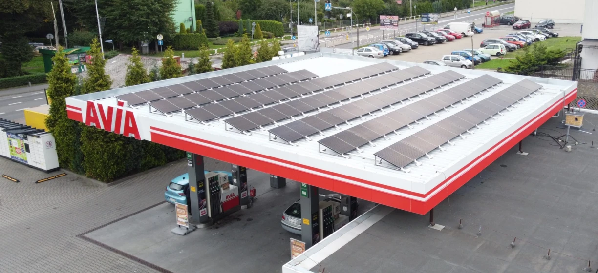 A car parked at an AVIA filling station with a bumper sticker and vehicle registration plate in a parking lot. Automotive mirror and fuel dispenser in the background.