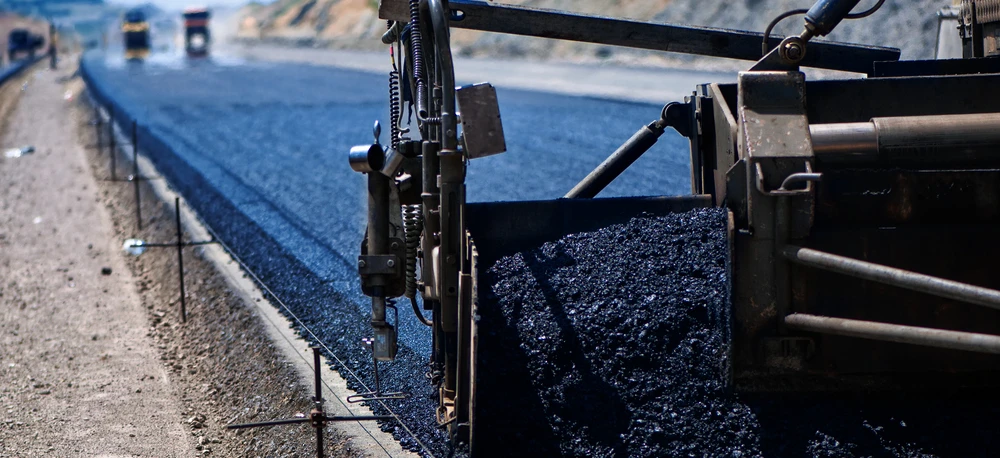 A machine lays asphalt on a road surface with heavy equipment and building materials nearby. Construction site with concrete, gravel, and tar.
