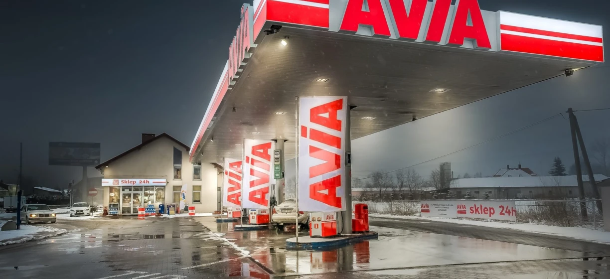 Cysternowa ciężarówka paliwowa zaparkowana na stacji benzynowej czynnej 24 godziny na dobę (Sklep 24h) z widocznym logo AVÍA na zderzaku.