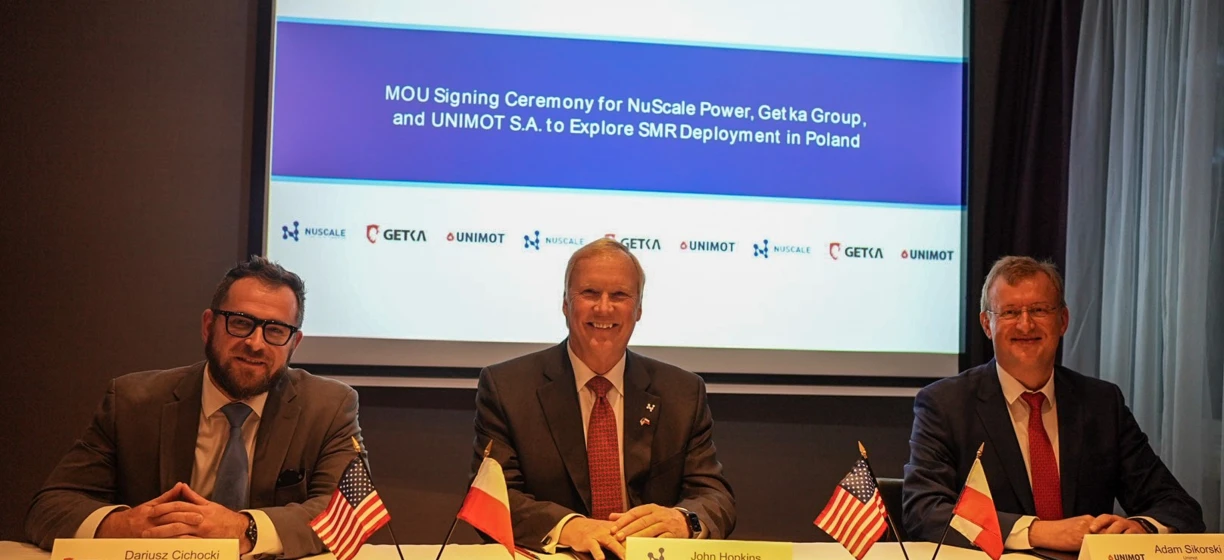 Image alt-text: CEOs of NuScale Power, Getka Group, and UNIMOT S.A. signing MOU at a ceremony in Poland. Electronic devices, suits, and microphones visible.