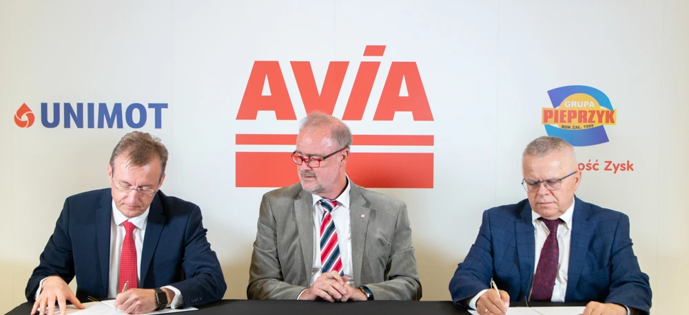 Alt-text: Professional white-collar worker in a suit and necktie, standing in front of an advertising display for AVIA company. Company logo and information in Polish language.