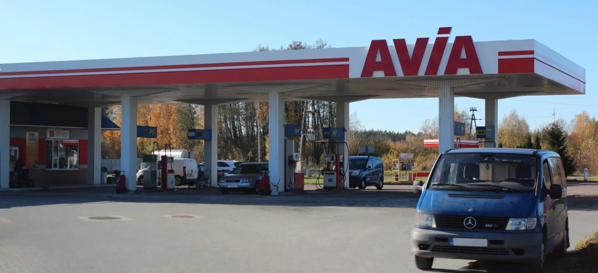 A family car at a gas station filling up with gasoline. The vehicle's bumper, mirrors, and registration plate are visible. Signs show the location as KIOPIST Sklep ON AVIA 94.