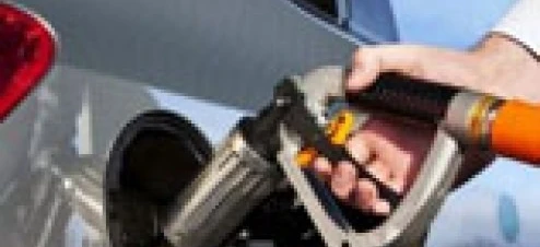 A fuel dispenser at a filling station next to a car with its door open and a windshield being cleaned. Automotive care with electric vehicle and hood in background.
