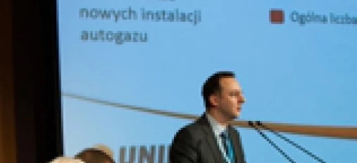A spokesperson in a formal suit and coat is giving a speech at an event using audio equipment and a microphone. Text on the image mentions the total number of LPG conversions in 2012.
"Spokesperson in formal wear at an event discussing LPG conversions in 2012. Audio equipment and microphone visible."