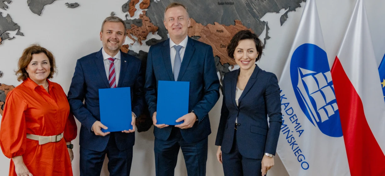 A man in a formal suit and blazer with a smile on his face standing in front of flags of Greenland, Russia, Mongolia, China, UNIMOT, KADEMIA, and MINSKIEGO universities.