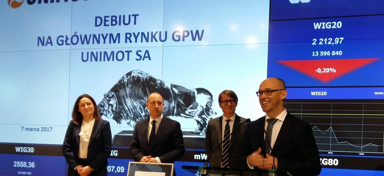 Alt-text: A professional in a formal white-collar worker attire with a coat, blazer, and logo of UNIMOT. Stock market data in Polish on the Warsaw Stock Exchange.