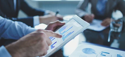 A group of businesspeople are sitting around a table having a meeting. The focus is on a white-collar worker gesturing whilst using a communication device.