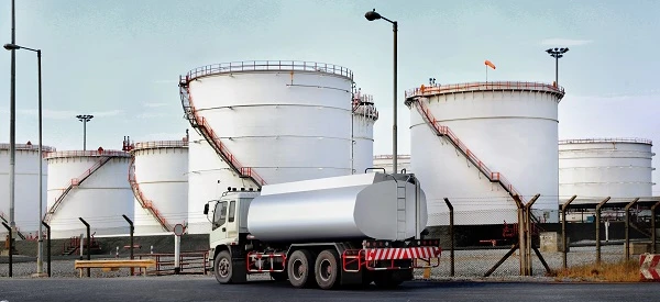 A commercial truck with a fuel tank parked next to a storage silo. A cylindrical machine with a pipe connected to it in an engineering setting.