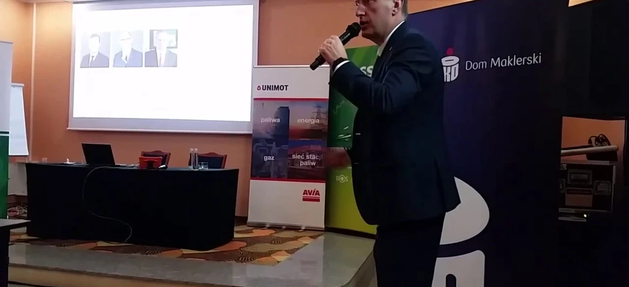 Alternatywny tekst: Mężczyzna w garniturze prezentujący na urządzeniu multimedialnym podczas wydarzenia. Słowa kluczowe: Urządzenie elektroniczne, Odzież wierzchnia, Prezentacja, Technologia, UNIMOT.