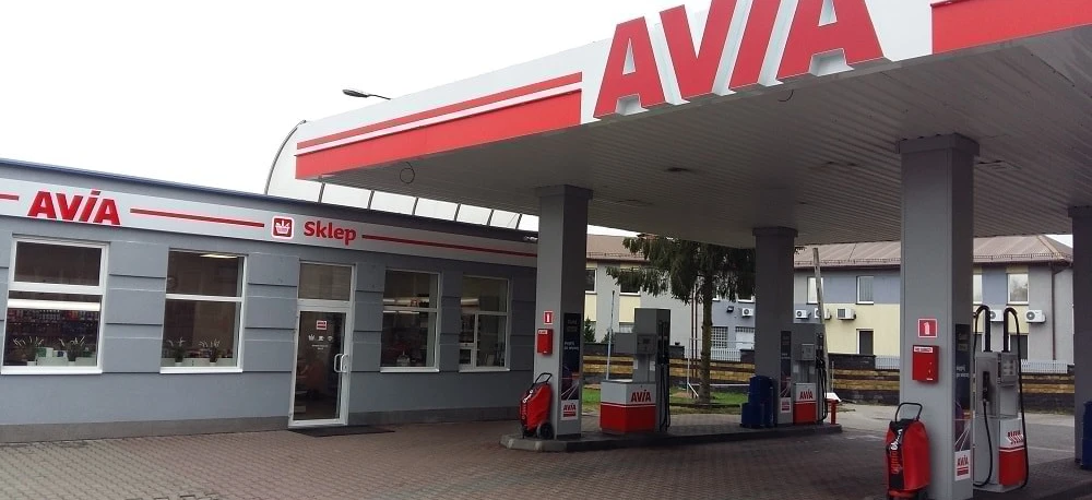 A close-up of a fuel dispenser at an AVIA filling station with an automotive mirror, bumper, and vehicle registration plate in the background. -AVIA gas station.