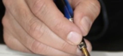A person's hand holding a pen over a piece of paper on a desk with office supplies. Writing with a wrist resting on the table.