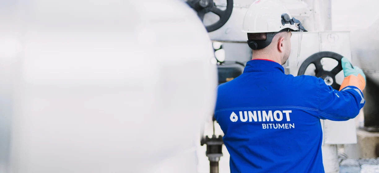 Robotnik fizyczny noszący odzież roboczą w fabryce, oznakowanej UNIMOT BITUMEN. Praca dla inżyniera lub rzemieślnika.