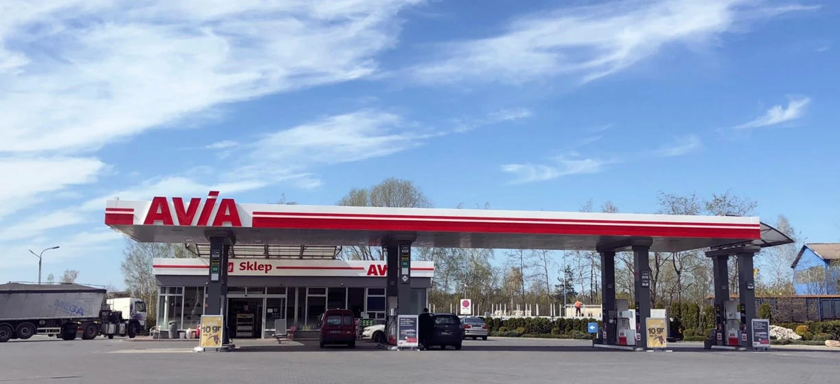 A close-up view of a family car parked at a filling station, showing the bumper, side-view mirror, and car door. The image also features a sport utility vehicle and a subcompact car in the background.