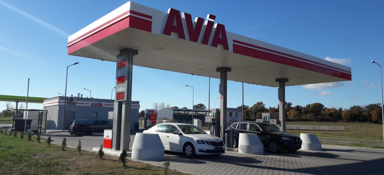 Alt-text: A fuel pump displaying prices for gasoline and diesel, with reflection of a subcompact car in the automotive side-view mirror. Bright colors of blue and red at the filling station with AVIA signage.