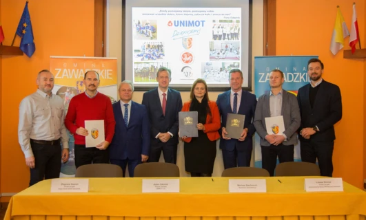 Grupa pracowników umysłowych w strojach formalnych, w tym garniturach, marynarkach i krawatach, stojąca i uśmiechająca się. Logo z tekstem "GMINA" widoczne.
