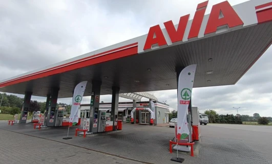 Image of a car parked at a filling station with the fuel pump nozzle in the gas tank. The vehicle's mirrors, bumper, and registration plate are visible.