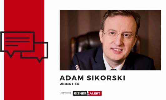 Profesjonalny mężczyzna w garniturze i okularach, zidentyfikowany jako Adam Sikorski z UNIMOT SA, w dyskusji biznesowej dla BIZNES ALERT.