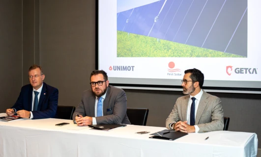 A white-collar worker in formal wear holding a microphone at an event featuring clothing, outerwear, and electronic devices. UNIMOT, GETKA, and First Solar logos featured prominently.