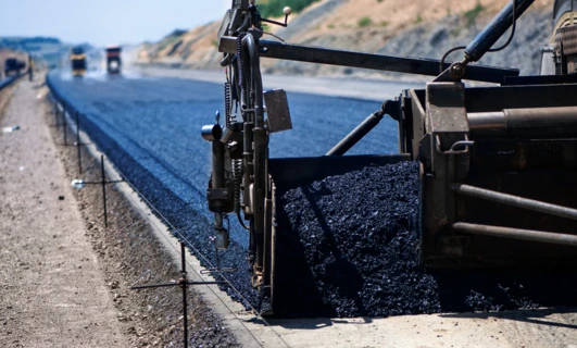 A machine lays asphalt on a road surface with heavy equipment and building materials nearby. Construction site with concrete, gravel, and tar.
