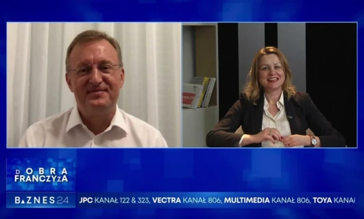 Pracownik umysłowy wygłaszający przemowę w programie telewizyjnym na temat możliwości biznesowych. Tekst na wyświetlaczu zawiera słowa kluczowe takie jak "żyza", "DOBRA", "FRANCZYZA", "BAZNES".