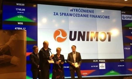 Pracownik umysłowy w garniturze patrzący na urządzenie wyświetlające informacje finansowe. Widoczny jest logo GPW i WIG20. Wyświetlane są dane dotyczące zarządzania i pracy.