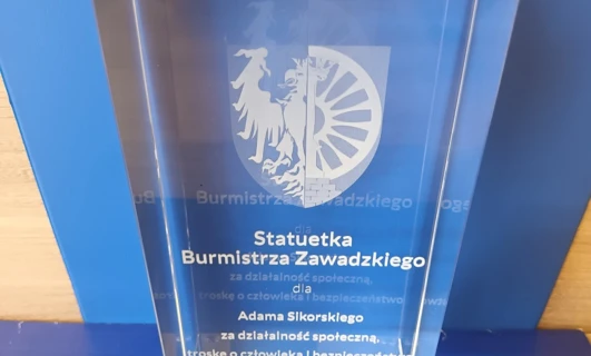 Obraz trofeum z napisem "Nagroda za działalność społeczną, troska o bezpieczeństwo ludzkie" wręczony przez burmistrza Zawadzkiego. Tło wykonane z transparentności i grafiki.