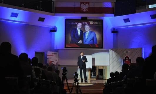 A spokesperson in a suit giving a speech at an event while standing at a podium with a display device and electronic devices in the background. Clothing includes a coat and outerwear. QUNIMOT 2016 presentation.