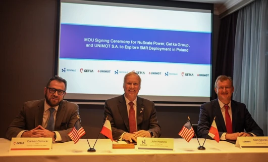 Image alt-text: CEOs of NuScale Power, Getka Group, and UNIMOT S.A. signing MOU at a ceremony in Poland. Electronic devices, suits, and microphones visible.