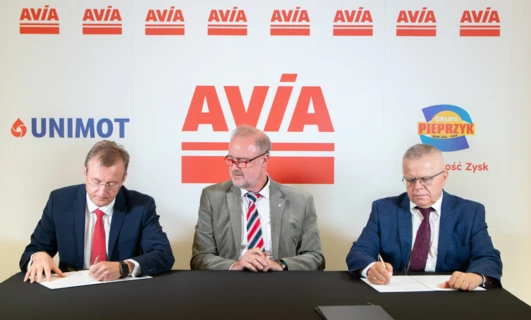 Tekst alternatywny: Profesjonalny pracownik umysłowy w garniturze i krawacie, stojący przed reklamowym panelem firmy AVIA. Logo firmy i informacje w języku polskim.