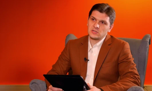 Profesjonalny biznesmen w marynarce i koszuli wygłaszający przemówienie, z pomarańczowym tłem i białym kołnierzykiem widocznym.