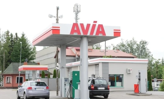 A family car parked at a fuel station with a fuel dispenser in the background. The vehicle's bumper, mirrors, and license plate are visible. AVIA and SD Eibar logos are also present.