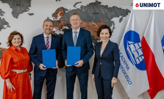 A man in a formal suit and blazer with a smile on his face standing in front of flags of Greenland, Russia, Mongolia, China, UNIMOT, KADEMIA, and MINSKIEGO universities.