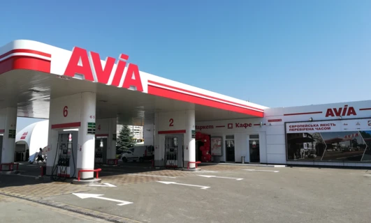 Alt-text: Automotive side-view mirror and bumper of a sport utility vehicle parked at a filling station. Image also shows rear-view mirror, hubcap, and car door with the AVIA brand logo.