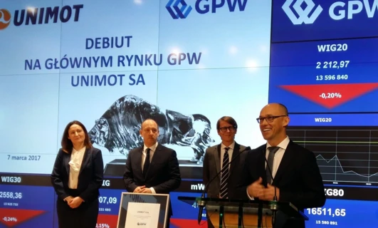 Alt-text: A professional in a formal white-collar worker attire with a coat, blazer, and logo of UNIMOT. Stock market data in Polish on the Warsaw Stock Exchange.