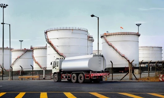 A commercial truck with a fuel tank parked next to a storage silo. A cylindrical machine with a pipe connected to it in an engineering setting.