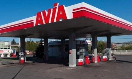 A car parked at a filling station with a shiny bumper and side-view mirror. Vehicle registration plate visible on the car door. AVIA logo on the hardtop.