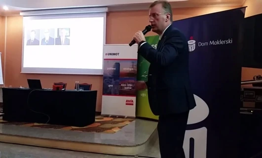 Alternatywny tekst: Mężczyzna w garniturze prezentujący na urządzeniu multimedialnym podczas wydarzenia. Słowa kluczowe: Urządzenie elektroniczne, Odzież wierzchnia, Prezentacja, Technologia, UNIMOT.