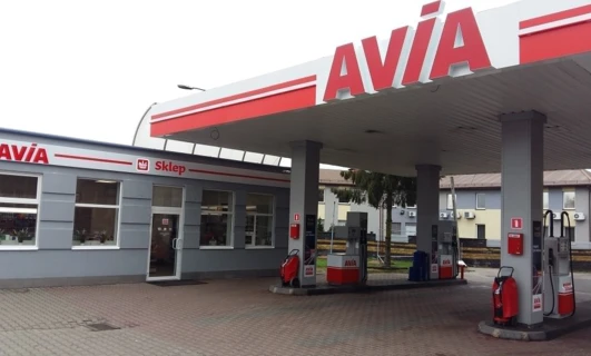 A close-up of a fuel dispenser at an AVIA filling station with an automotive mirror, bumper, and vehicle registration plate in the background. -AVIA gas station.