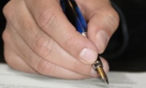 A person's hand holding a pen over a piece of paper on a desk with office supplies. Writing with a wrist resting on the table.