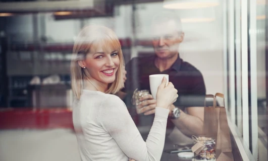 A woman with blond hair smiling while holding a coffee cup. Tableware and drinkware in focus, conveying happiness and enjoyment.