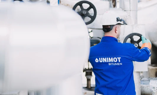 Robotnik fizyczny noszący odzież roboczą w fabryce, oznakowanej UNIMOT BITUMEN. Praca dla inżyniera lub rzemieślnika.