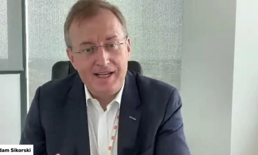 Adam Sikorski, pracownik umysłowy, nosi okulary i koszulę. Jego czoło i podbródek są widoczne powyżej kołnierza, gdy mówi jako rzecznik.