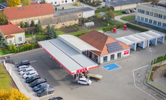 Ruchliwa stacja benzynowa z różnymi pojazdami zaparkowanymi, tankującymi paliwo pod jasnymi światłami parkingowymi i hamulcowymi. Zawiera samochody, ciężarówki i SUV-y z dystrybutorem w tle.