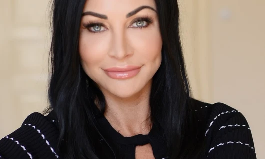 A smiling woman with long black hair, perfectly shaped eyebrows, full lips, and stunning eyelashes. Her beautifully layered hair is styled in a step cut, enhancing her beauty.
