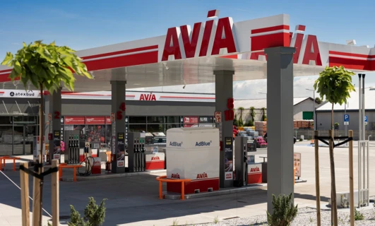 Obraz stacji tankowania paliwa z cysterną i SUV-em tankującymi benzynę. Logo Atexbud widoczne na dystrybutorze paliwa.