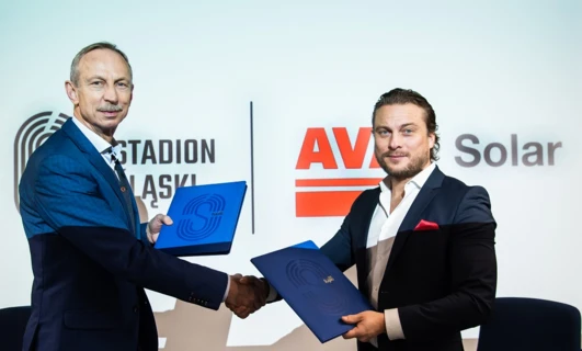 Pracownik umysłowy w eleganckim garniturze i marynarce uśmiecha się obok logo reklamowego AV Solar na STADIONIE. Promując profesjonalny styl i czyste rozwiązania energetyczne.