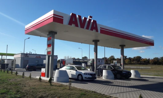 Alt-text: A fuel pump displaying prices for gasoline and diesel, with reflection of a subcompact car in the automotive side-view mirror. Bright colors of blue and red at the filling station with AVIA signage.