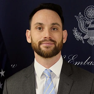 Mężczyzna z brodą i uśmiechem, ubrany w formalny strój, patrzący prosto w kamerę. Jego brwi są uniesione w przyjaznym wyrazie.